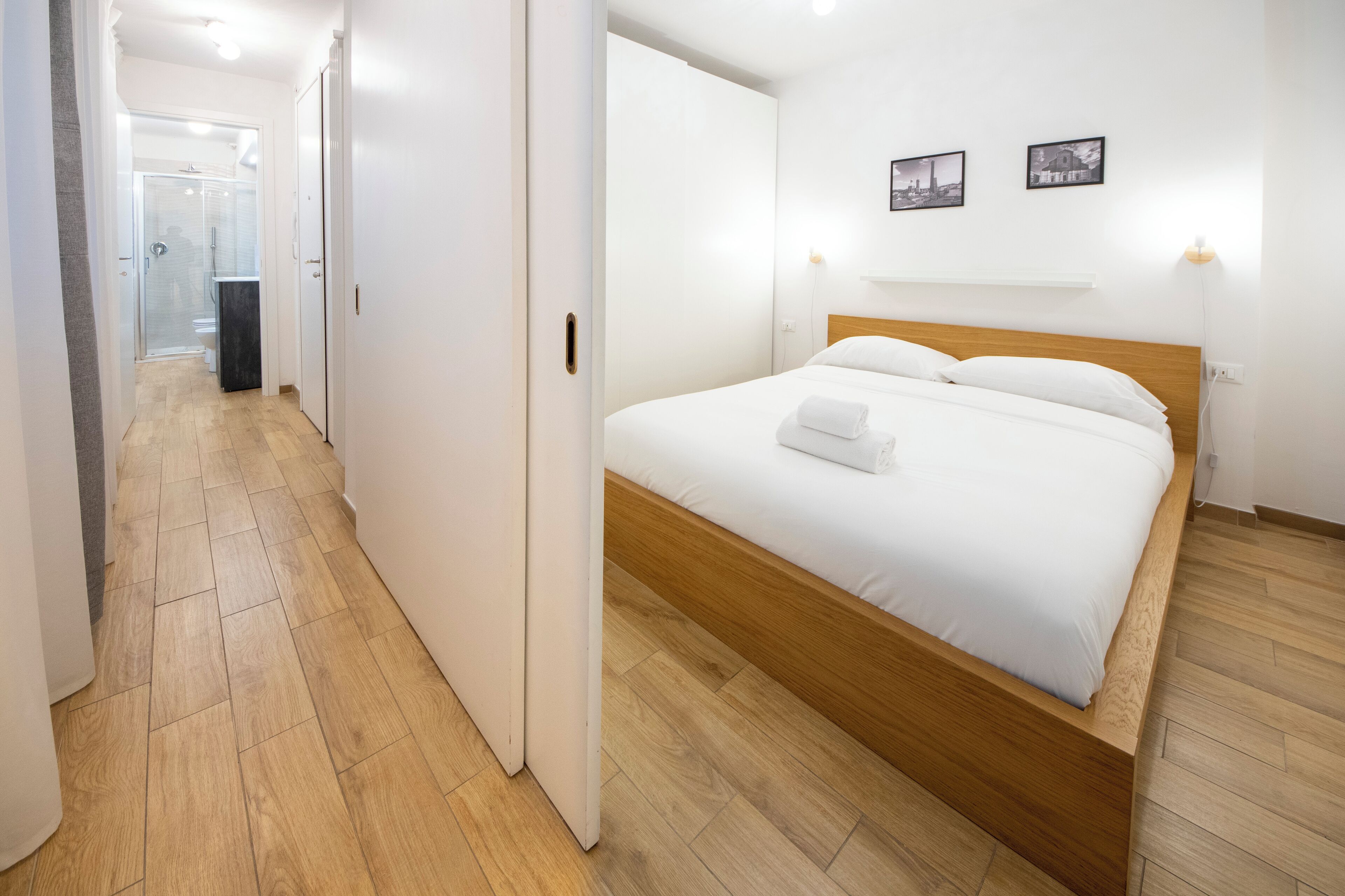 A hotel room with a wooden bed, white bedding, a glass shower, and a hallway with wooden flooring.