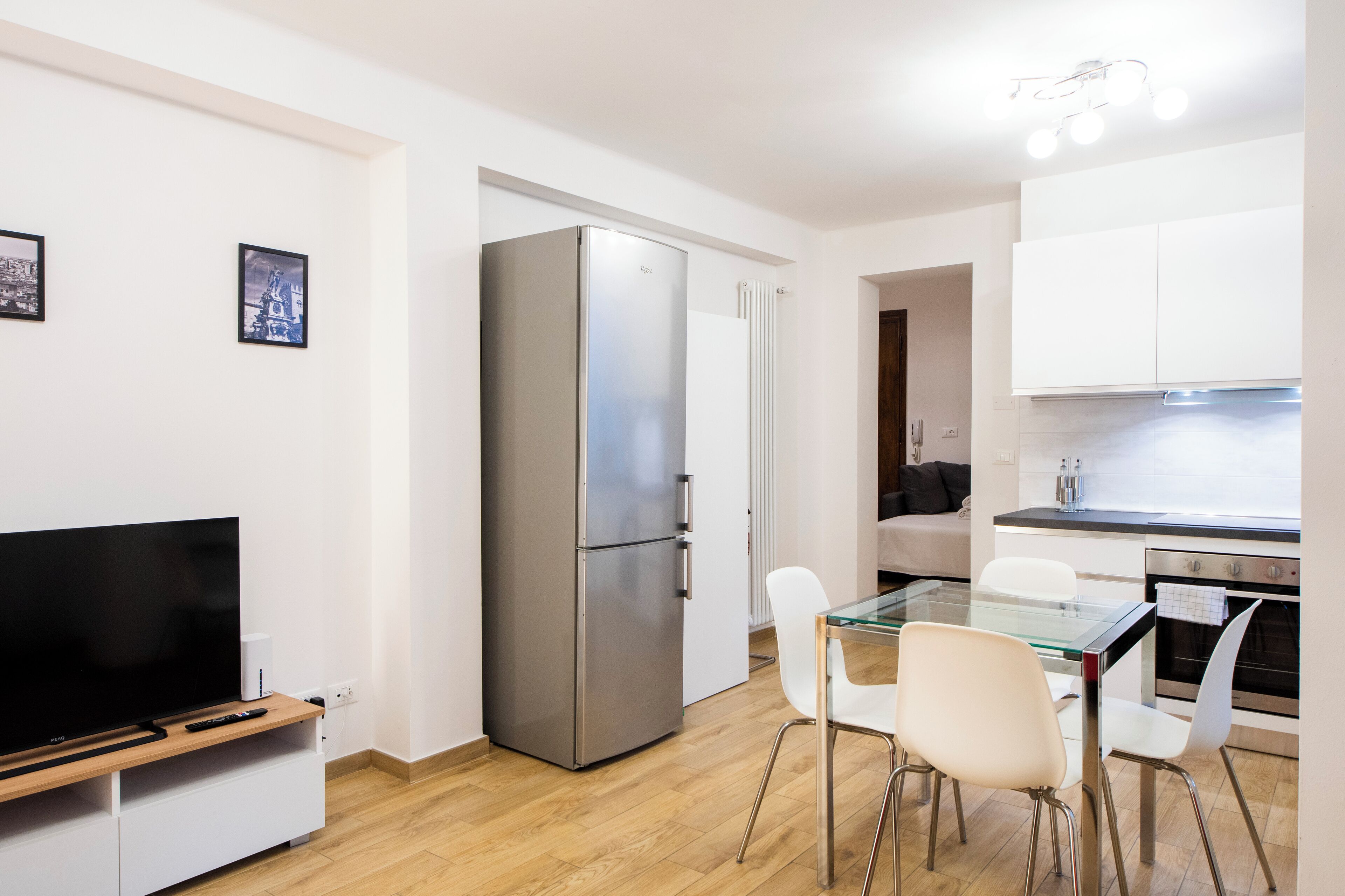 A compact kitchen with a refrigerator, a dining table with chairs, a TV on a stand, and a sofa in the adjacent living area.