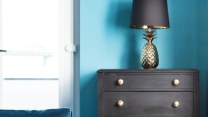 A room with a blue sofa, a dark wooden dresser with gold knobs, and a lamp with a black shade. - We Love HU -Sky Marina Suite (Kingston upon Hull)