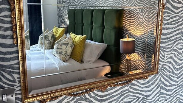 Un dormitorio con pared estampada de cebra, cabecero tapizado en verde y espejo con marco dorado.