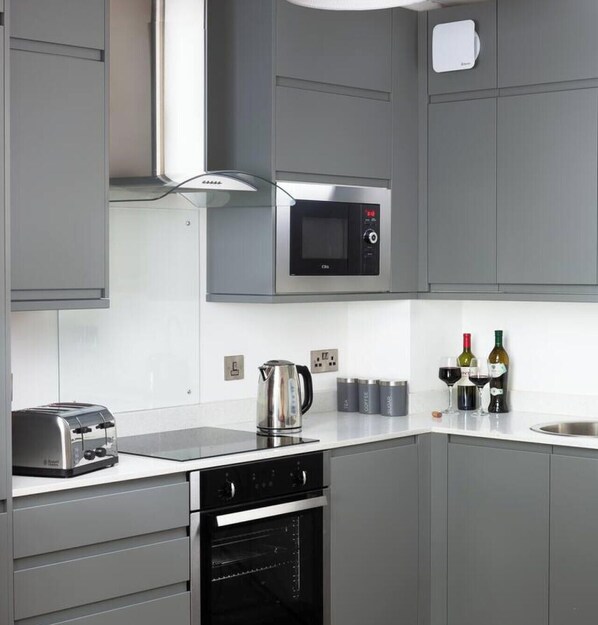 Une cuisine moderne avec des meubles gris, un four à micro-ondes encastré, un grille-pain, un four et une bouilloire. On peut voir des bouteilles de vin et des verres sur le plan de travail.