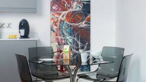 Un comedor moderno con una mesa de vidrio y sillas negras, un cuadro abstracto grande en la pared y una cafetera en la encimera.