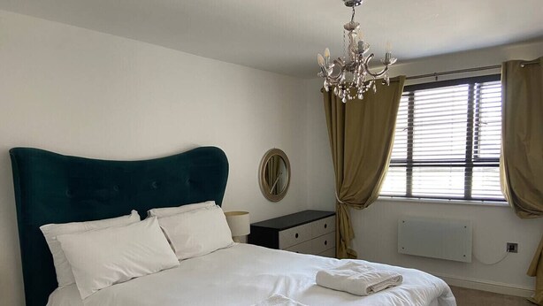 Una cama bien hecha con sábanas blancas y dos toallas dobladas, un cabecero tapizado en color verde oscuro, un espejo redondo en la pared, un candelabro y una ventana con cortinas.