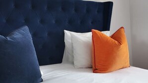 Una cama con cabecera azul oscuro, ropa de cama blanca y tres almohadas en azul, blanco y naranja.