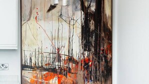 Un comedor con una mesa de vidrio y sillas de metal, un cuadro abstracto grande en la pared, y una lámpara colgante.