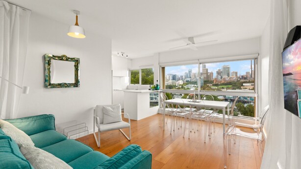 A modern living room with a teal sofa, a dining area with clear chairs, a city view through large windows, and a white ceiling fan. - Cosy 1-bed With Swimming Pool Facing Opera House (Sydney)