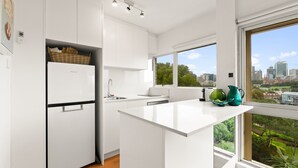 A modern kitchen with white cabinetry, a built-in refrigerator, a sink, and a large window offering a city view. - Cosy 1-bed With Swimming Pool Facing Opera House (Sydney)