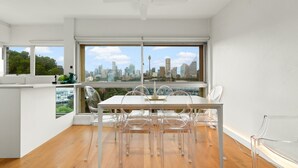 A modern kitchen with a large window offering a city view, a dining area with clear chairs, and a ceiling fan. - Cosy 1-bed With Swimming Pool Facing Opera House (Sydney)