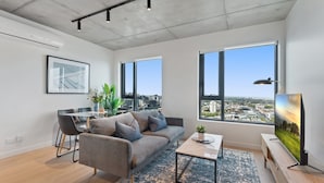 A modern living room with a gray sofa, a coffee table, and a TV on a wooden stand. There are large windows offering a city view, and a dining area with a table and chairs. - Sophisticated 2-bed With Bay Views & Amenities (Melbourne)