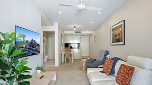 A modern living room with a white sofa, a grey armchair, and a TV displaying a cityscape. A ceiling fan and recessed lighting are visible. - Cosy 1-bed With River Views, Pool, Gym & BBQ (Brisbane)
