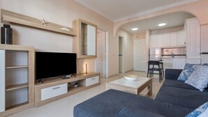 A modern living room with a flat-screen TV, a grey sofa, a coffee table, and a kitchen area with white cabinets.