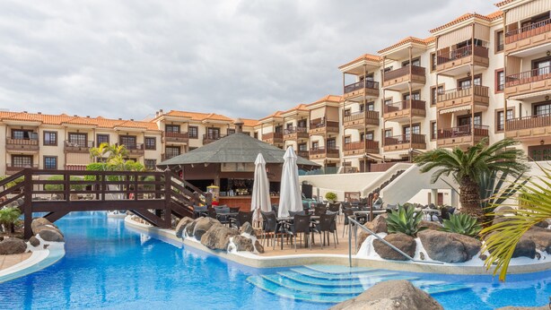 A swimming pool with a wooden bridge, surrounded by a resort building with balconies and outdoor seating.