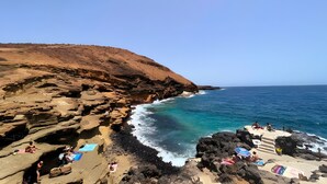 A rocky coastline with clear blue sea, people sunbathing, and a clear sky.
