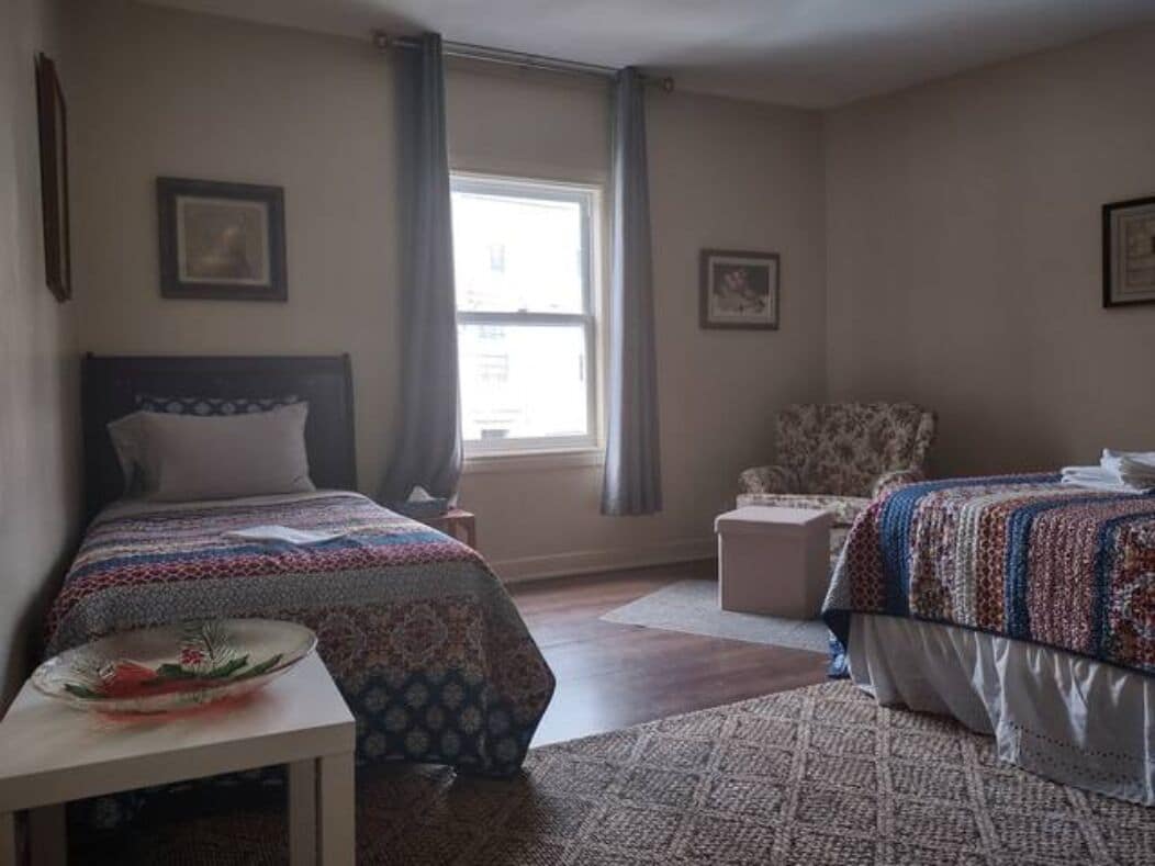 A bedroom with two beds, a window with curtains, a chair, and a small table with a bowl.