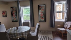 Un área de comedor con una mesa redonda, sillas y un tapete. Hay dos ventanas con cortinas, y obras de arte en las paredes.