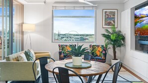 Un salon moderne avec une table ronde en bois, deux fauteuils colorés, un canapé, un ventilateur de plafond, une grande fenêtre donnant sur une grande roue et une plante en pot.