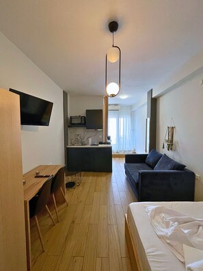 A modern hotel room with a wooden desk, a bed, a sofa, a TV mounted on the wall, a kitchenette, and a large window with curtains.