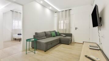 Een moderne woonkamer met een hoekbank, een klein groen bijzettafeltje, een flatscreen televisie aan de muur bevestigd en een houten bureau met een afstandsbediening en een tablet.