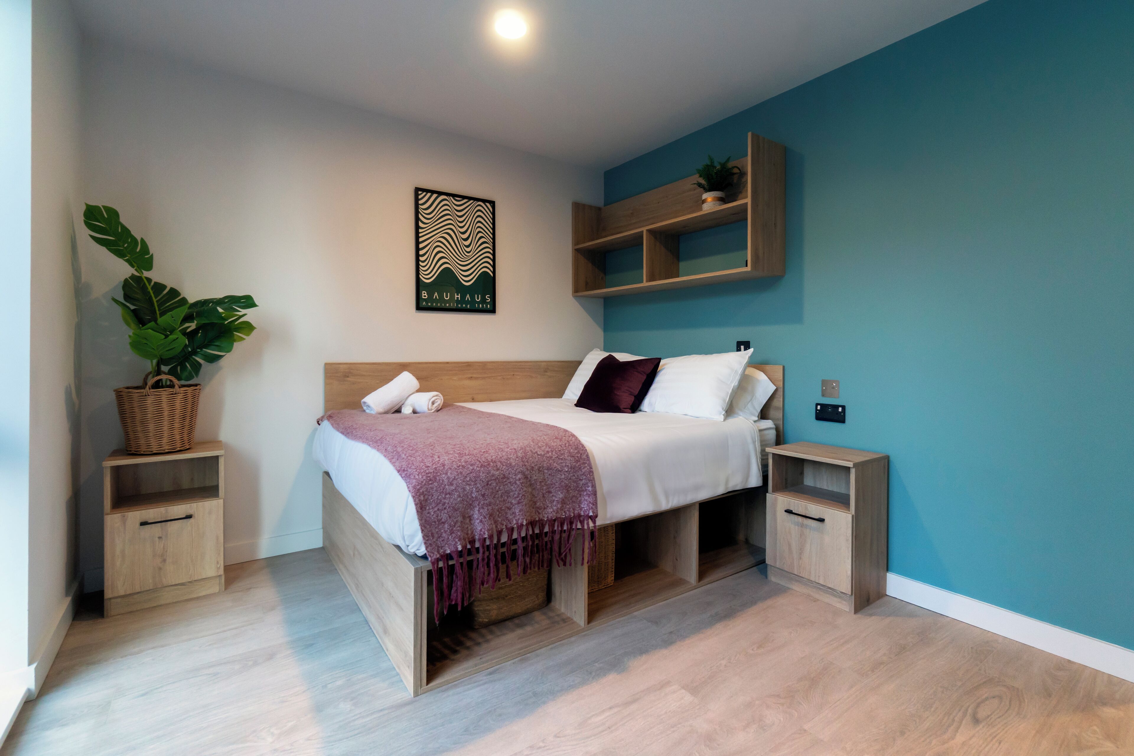 A modern bedroom with a wooden bed, a headboard, a wall-mounted shelf with a plant, and a framed poster.