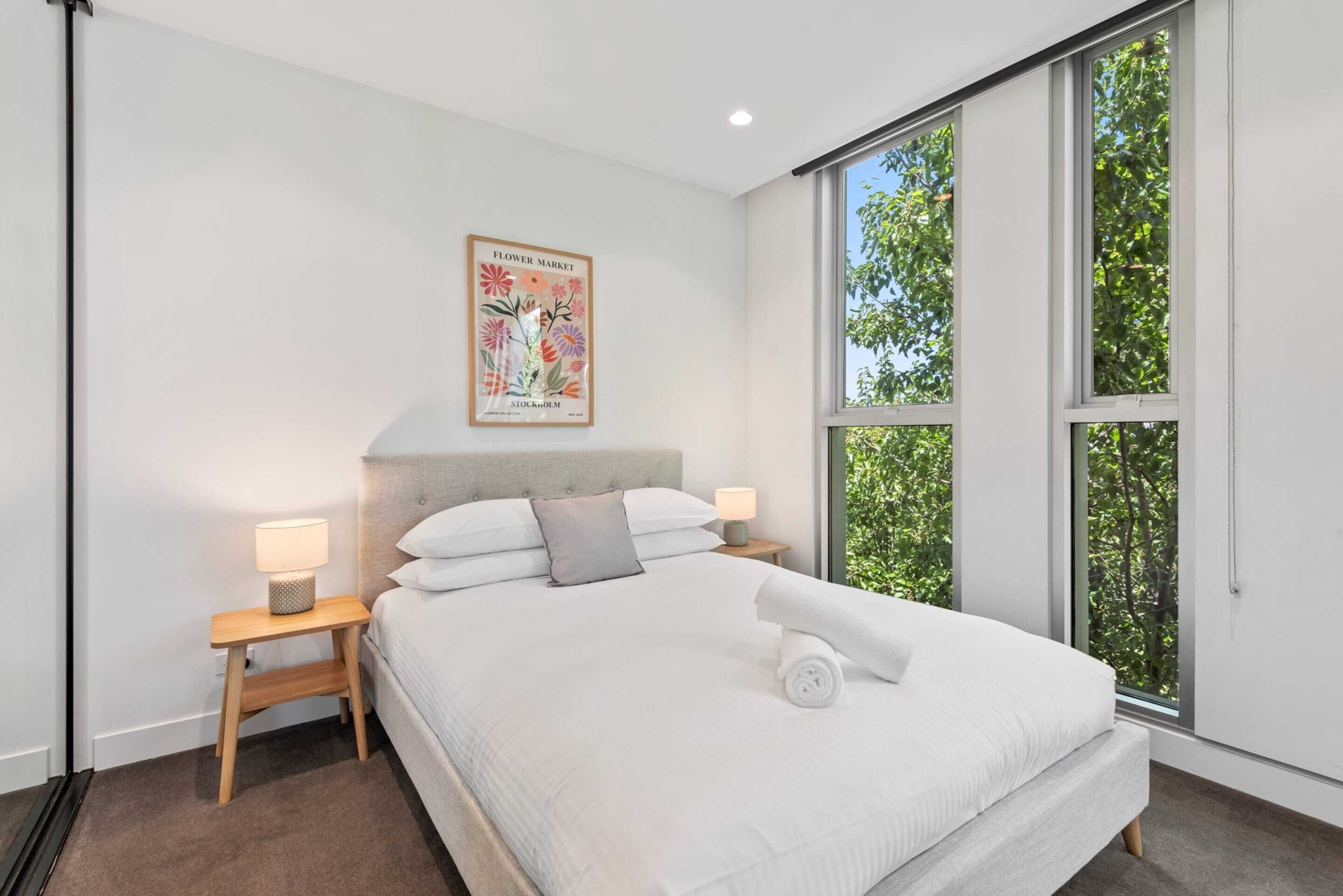 Un dormitorio moderno con una cama grande, dos mesitas de noche con lámparas, un cartel en la pared y vistas a la vegetación a través de amplias ventanas.