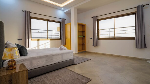 A modern bedroom with a bed, bedside table, lamp, and large windows. - Residence fakir (Dakhla)