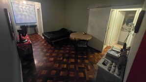 Een kamer met een houten vloer met een patroon, een kleine ronde tafel met stoelen, een zwarte bank, een scherm voor een beamer en een spiegel aan de muur.