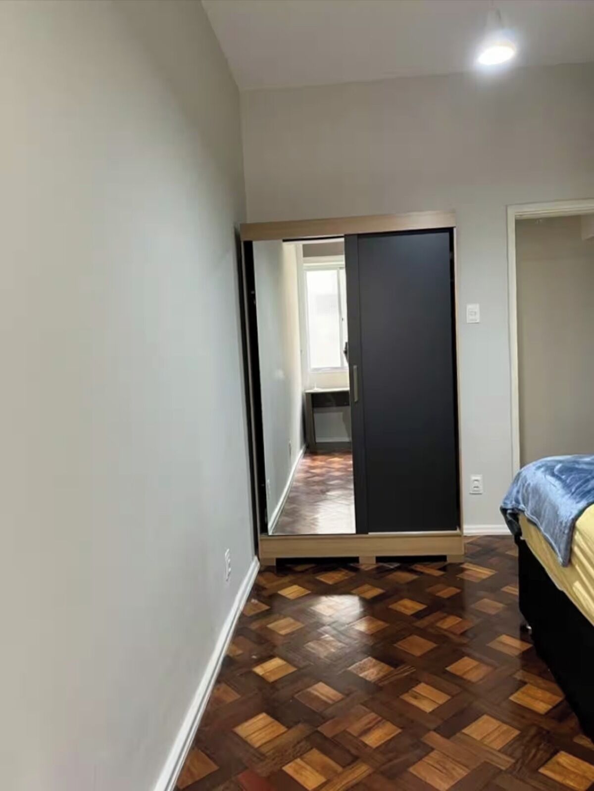 A bedroom with a wooden parquet floor, a bed with a blue cover, a wardrobe, and a mirror.