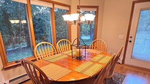 Une salle à manger avec une table et des chaises en bois, un lustre et de grandes fenêtres donnant sur la forêt.