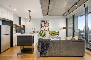 A modern kitchen with a central island, a grey sofa with cushions, and a large window offering a city view. - Modern, Executive 1 Bedroom Apartment With Balcony (Melbourne)