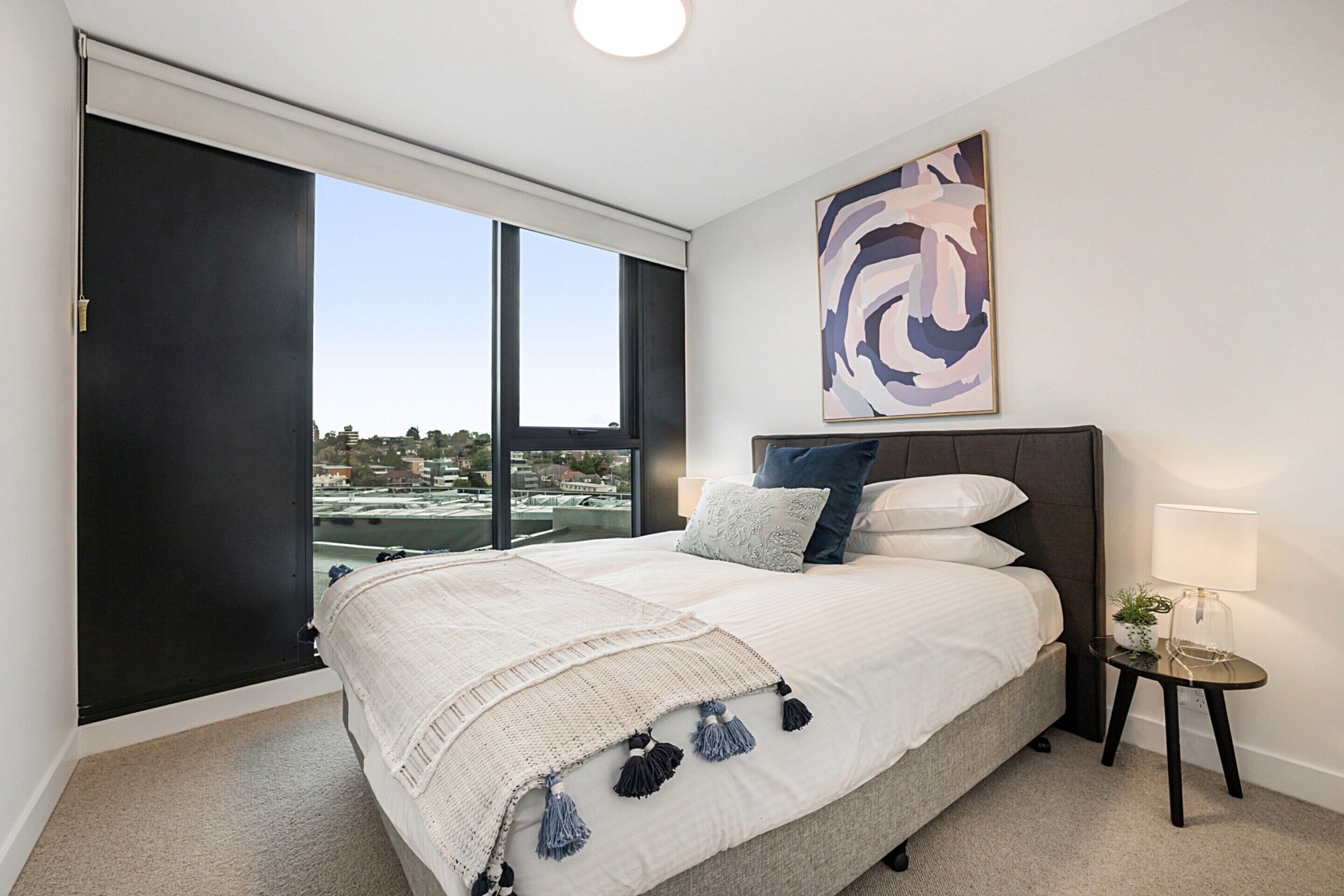 A bedroom with a large bed, a view through a sliding door, a wall-mounted abstract painting, and a small bedside table with a lamp.