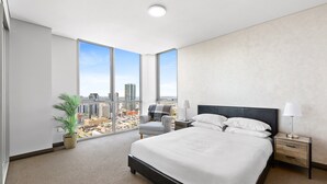 A modern hotel room with a large bed, a nightstand, a chair, and a view of the cityscape through large windows. - Central 2-bed With Parking & Stunning Views (Sydney)