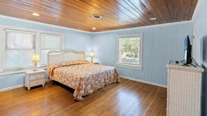 A bedroom with a wooden ceiling, a bed with a floral bedspread, two bedside lamps, a television on a stand, and a window with blinds. - A Block Away- 5 BR/3BA- Dog Friendly- Pool (North Myrtle Beach)