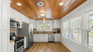 A kitchen with white cabinets, a wooden ceiling, and a view of the outdoors through large windows. - A Block Away- 5 BR/3BA- Dog Friendly- Pool (North Myrtle Beach)