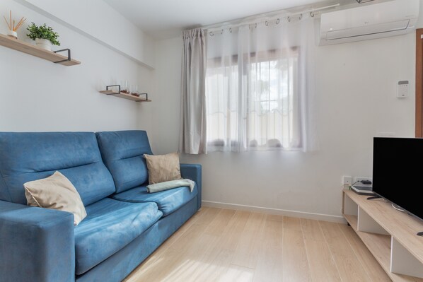 Un salón moderno con un sofá azul, un televisor de pantalla plana, suelo de madera y una ventana con cortinas traslúcidas.