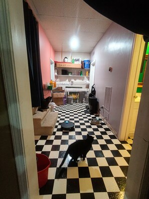 A narrow hallway with a checkered floor leading to a kitchen area with a refrigerator, microwave, and sink. A cat is visible on the floor, and there is a red bucket on the left side.