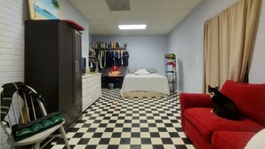 Una habitación con suelo a cuadros, un sofá rojo, una cama con manta blanca, un armario, un perchero y un cuadro de palmeras.