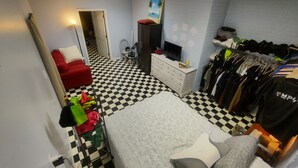 Una habitación con piso a cuadros, una cama con almohadas, una silla roja, un televisor sobre una cómoda y un perchero.