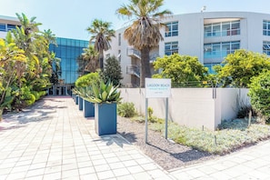 Une enseigne indiquant l’emplacement des appartements Lagoon Beach à Le Cap, avec un bâtiment moderne et un chemin pavé bordé de plantes.