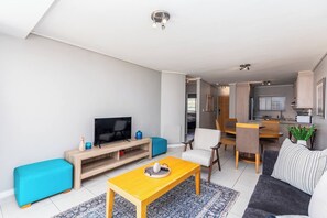 Una sala moderna con un televisor de pantalla plana, una mesa de centro de madera y un sofá. Hay una zona de comedor con una mesa y sillas de madera. Se ve la cocina al fondo, con electrodomésticos modernos.