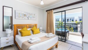 Une chambre d’hôtel avec un lit, une table de chevet, une lampe, un tableau, une fenêtre avec des rideaux et une vue sur une piscine et des bâtiments.