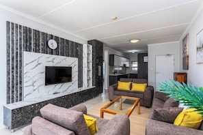 A modern living room with a flat-screen TV, grey sofas with yellow cushions, a marble accent wall, and a glass coffee table.