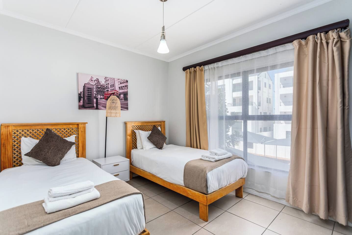 Ein Hotelzimmer mit zwei Betten, einem Nachttisch, einer Lampe, einem Fenster mit Vorhängen und Blick auf die Stadt.