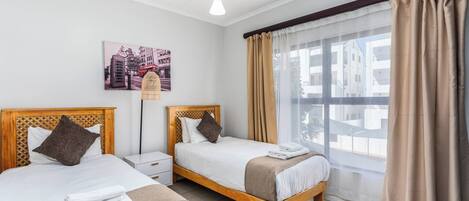 A hotel room with two beds, a nightstand, a lamp, a window with curtains, and a cityscape view.