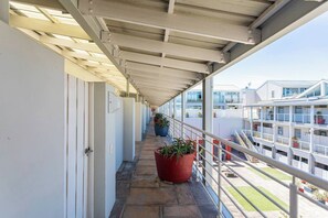 A covered balcony with potted plants, a railing, and a view of buildings in the background.