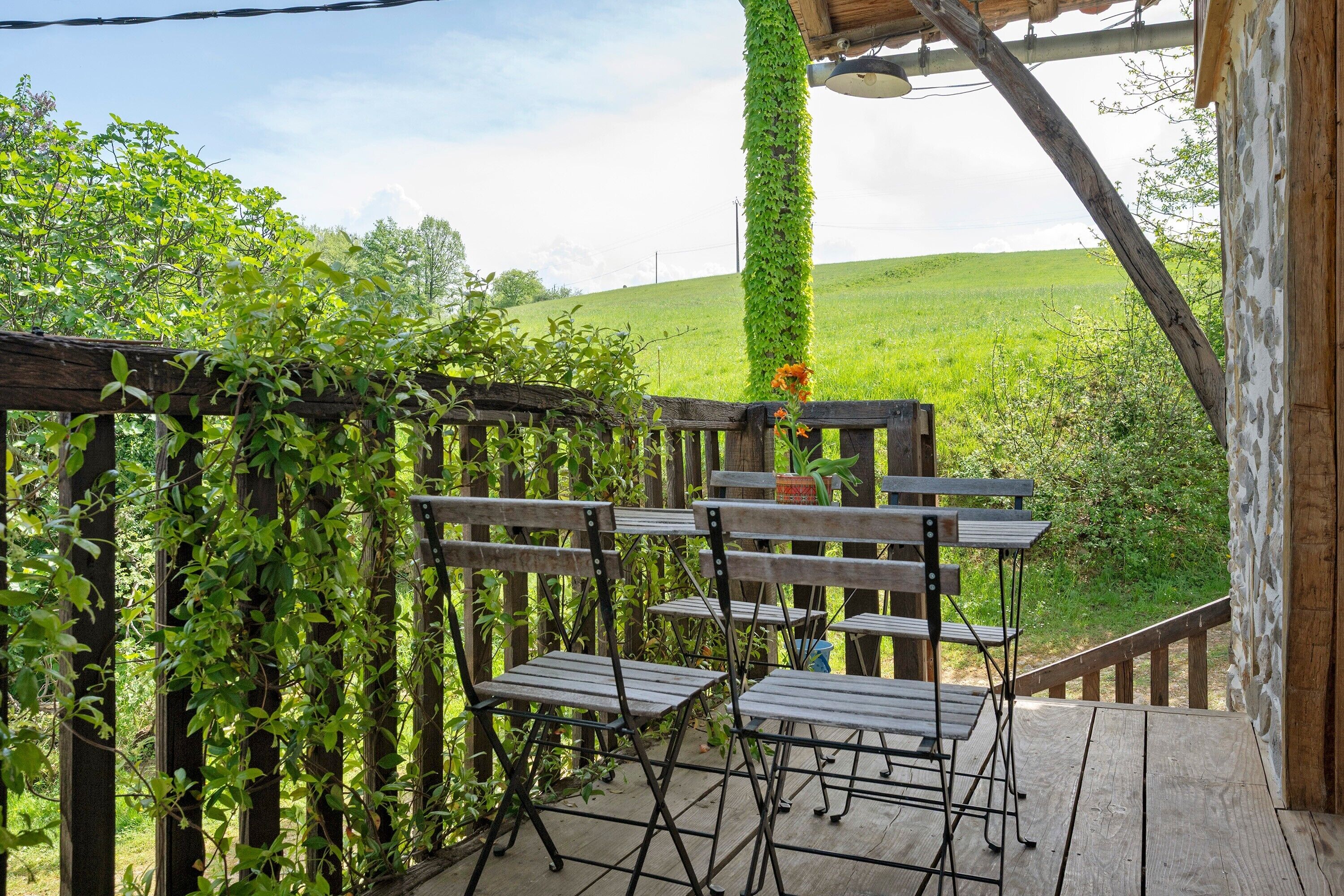 A wooden deck with metal chairs, surrounded by greenery and a view of a grassy field.