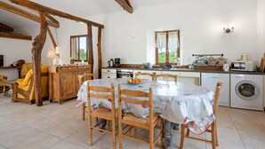 Una cuina amb taula de dinar, caires, armaris de fusta, rentadora i una finestra amb cortines.