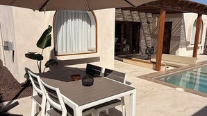 Um pátio externo com mesa, cadeiras, um grande guarda-sol e uma piscina.