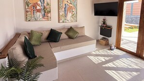 Sala de estar com teto de madeira, sofá com almofadas, duas obras de arte emolduradas, uma televisão sobre suporte preto e um vaso com planta.