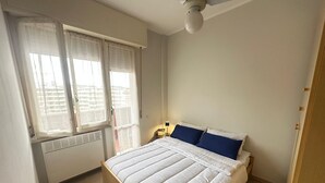 A bedroom with a bed, a window with blinds, a ceiling fan, and a nightstand with a lamp. - 'Between Sky and Sea by Vale' Apartment with Balcony and Air Conditioning (Fano)