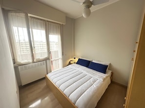 A bedroom with a bed, a window with blinds, a ceiling fan, and a nightstand with a lamp.